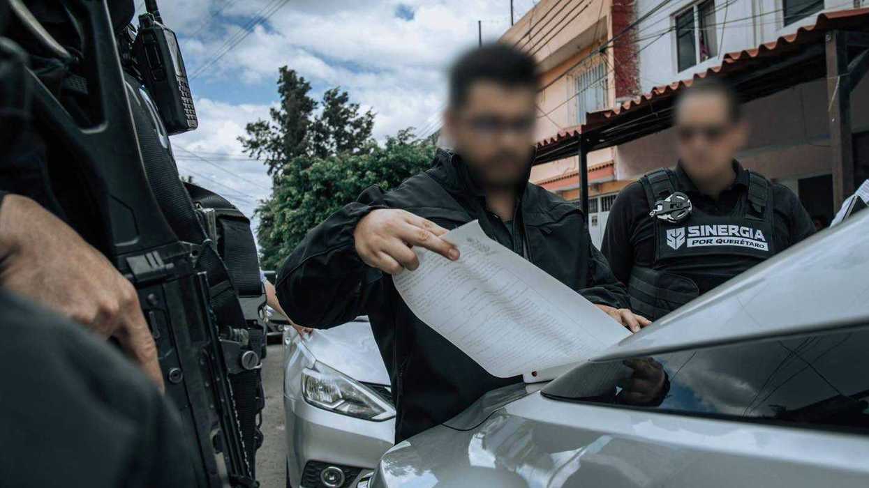 Agente de la Fiscalía de Querétaro revisa un documento sobre el cofre de un vehículo junto a elementos de Sinergia por Querétaro, con rostros difuminados.