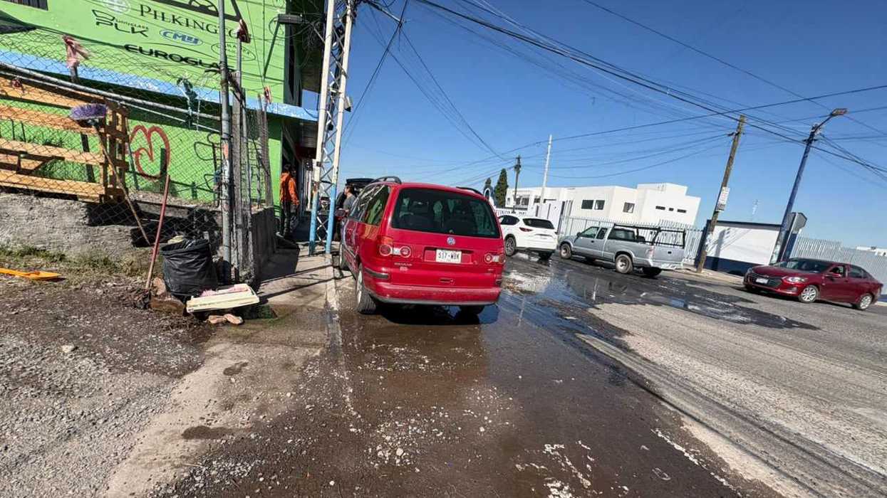 Agua de una fuga escurre sobre el pavimento de avenida Río Moctezuma, en San Juan del Río, junto a vehículos estacionados y en circulación.
