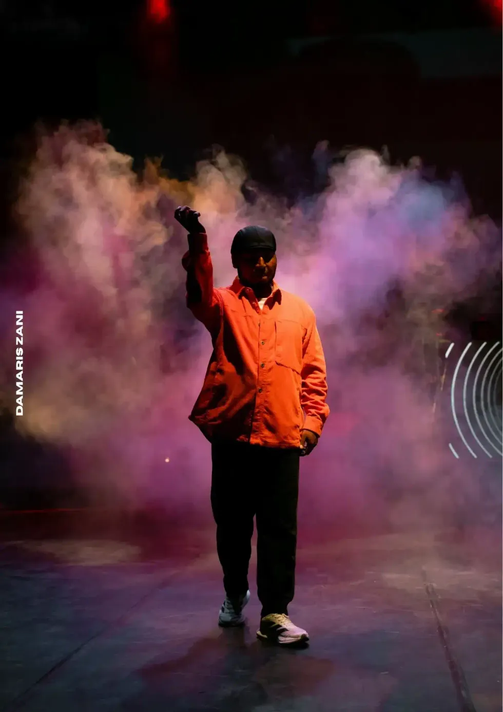 Artista con camisa naranja sobre un escenario cubierto por humo de colores durante una presentaci\u00f3n de cultura urbana.