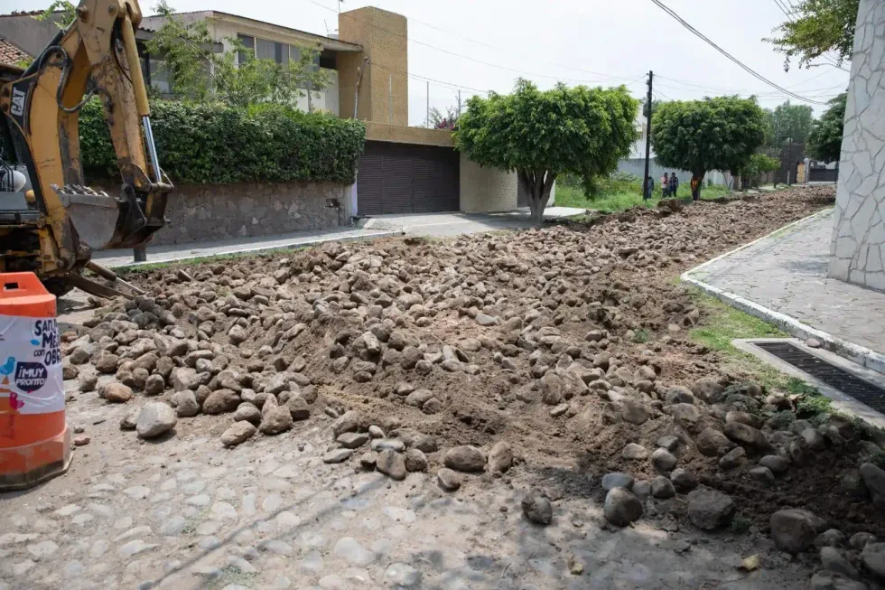 Calle con empedrado levantado y mont\u00edculos de piedra junto a una excavadora en una zona residencial