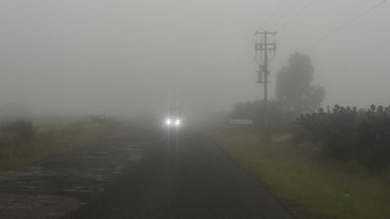 Camino rural cubierto de niebla densa, con un vehículo de frente con los faros encendidos y un poste de luz junto a la carretera.
