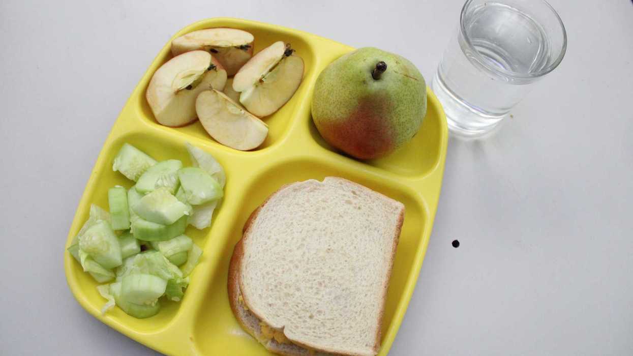 Charola escolar amarilla con sándwich de pan integral, manzana rebanada, pera y pepino, más vaso de agua.