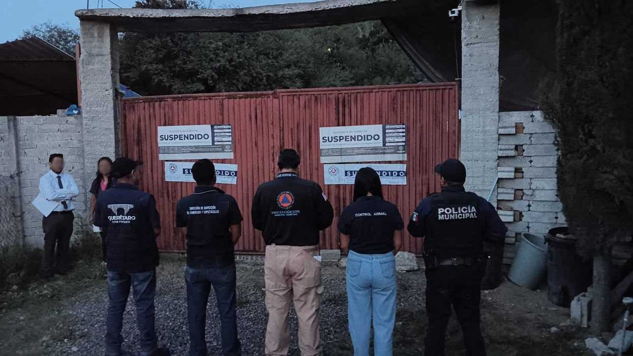 Cinco funcionarios municipales, de espaldas, frente a un portón metálico rojo con sellos de suspensión, al caer la tarde.