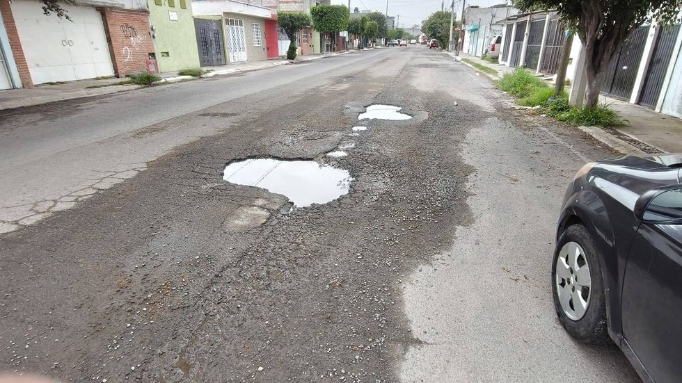 Cruce de calles con varios baches llenos de agua turbia y asfalto fracturado junto a una jardinera de ladrillo