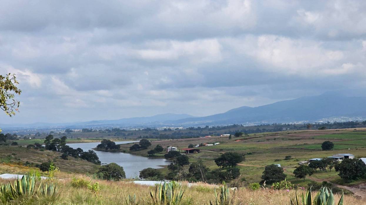 Cuerpo de agua entre lomeríos y campos de cultivo, con montañas al fondo bajo un cielo nublado