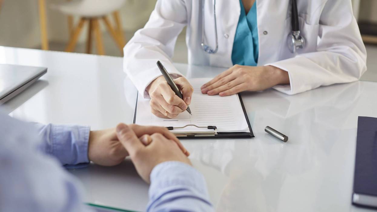 Doctora con bata blanca y estetoscopio escribe en un portapapeles durante una consulta médica con un paciente