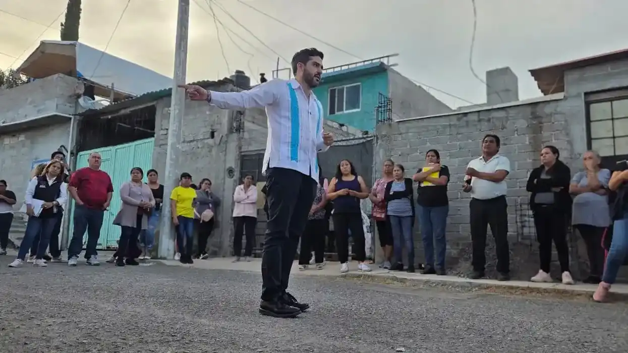 Dorantes Lámbarri señala con el brazo extendido frente a residentes del asentamiento Los Pinos reunidos en la calle, con viviendas de block y cielo nublado al fondo.