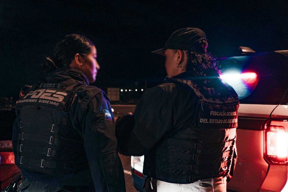 Dos agentes de seguridad, vistas de espaldas, junto a una patrulla con la torreta encendida durante un operativo nocturno; uno porta chaleco de la Polic\u00eda Estatal de Quer\u00e9taro y otro de la Fiscal\u00eda General del Estado de Quer\u00e9taro.