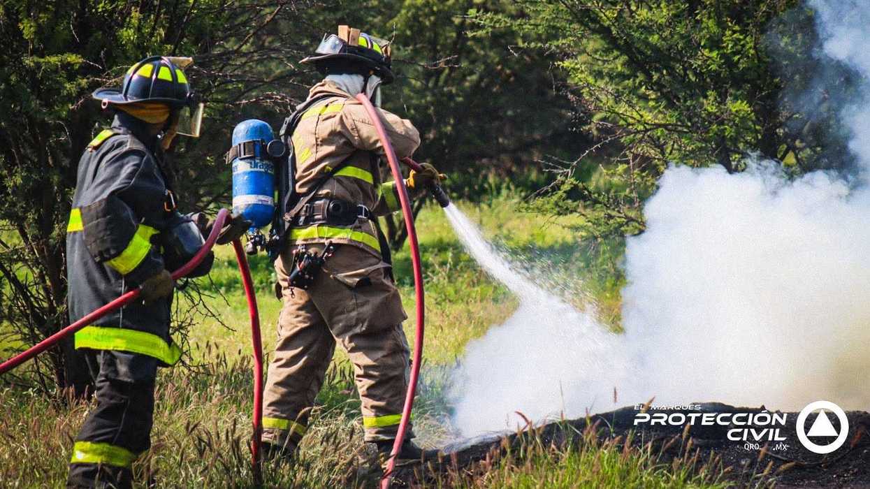 Dos bomberos de Protección Civil El Marqués, con equipo completo de protección, aplican agua con manguera sobre un foco de humo denso durante el control de un incendio de llantas en zona de vegetación en Santa Fe Libertadores.