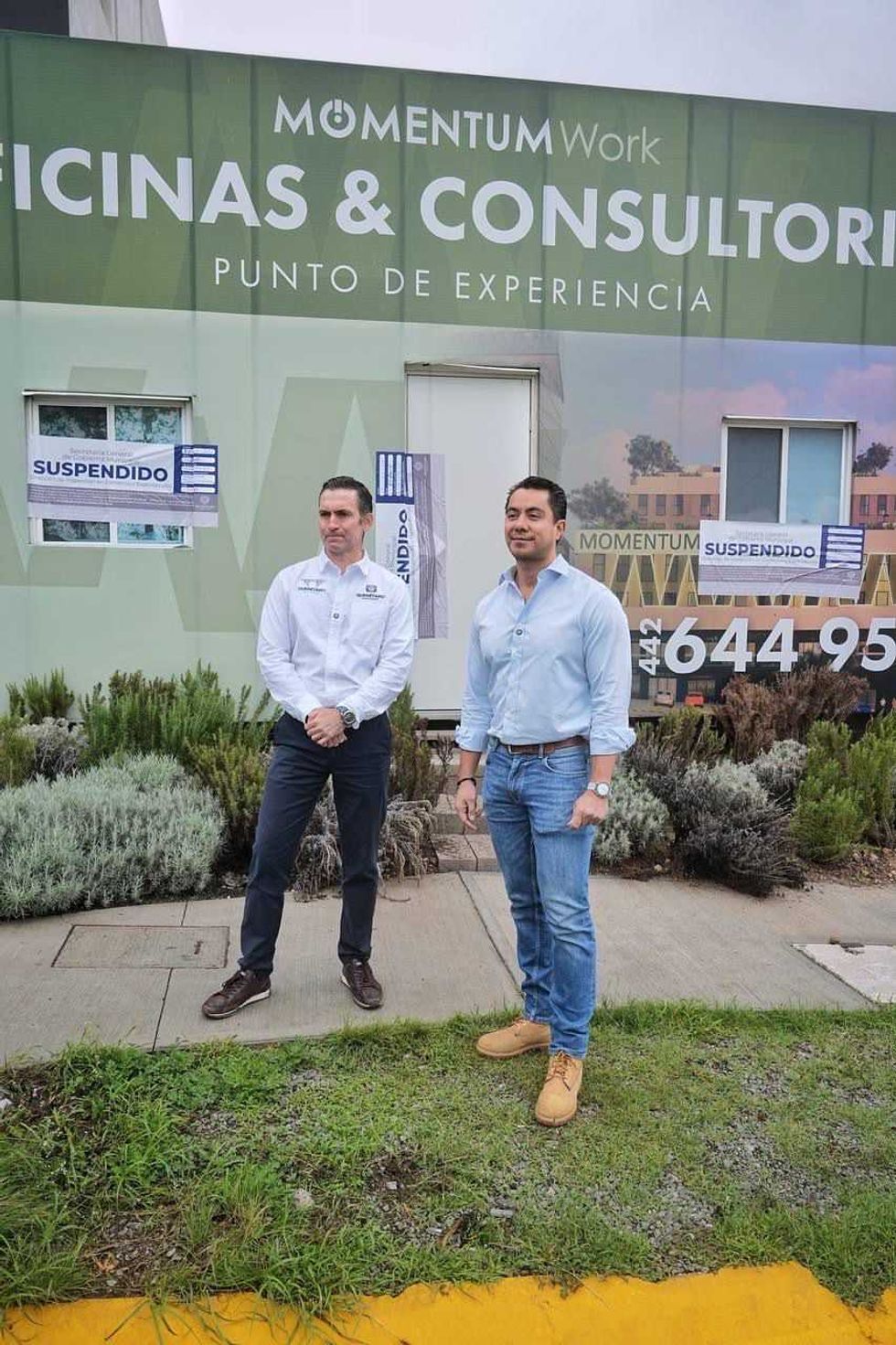 Dos funcionarios posan frente a la fachada del desarrollo Momentum Work con sellos de suspensi\u00f3n