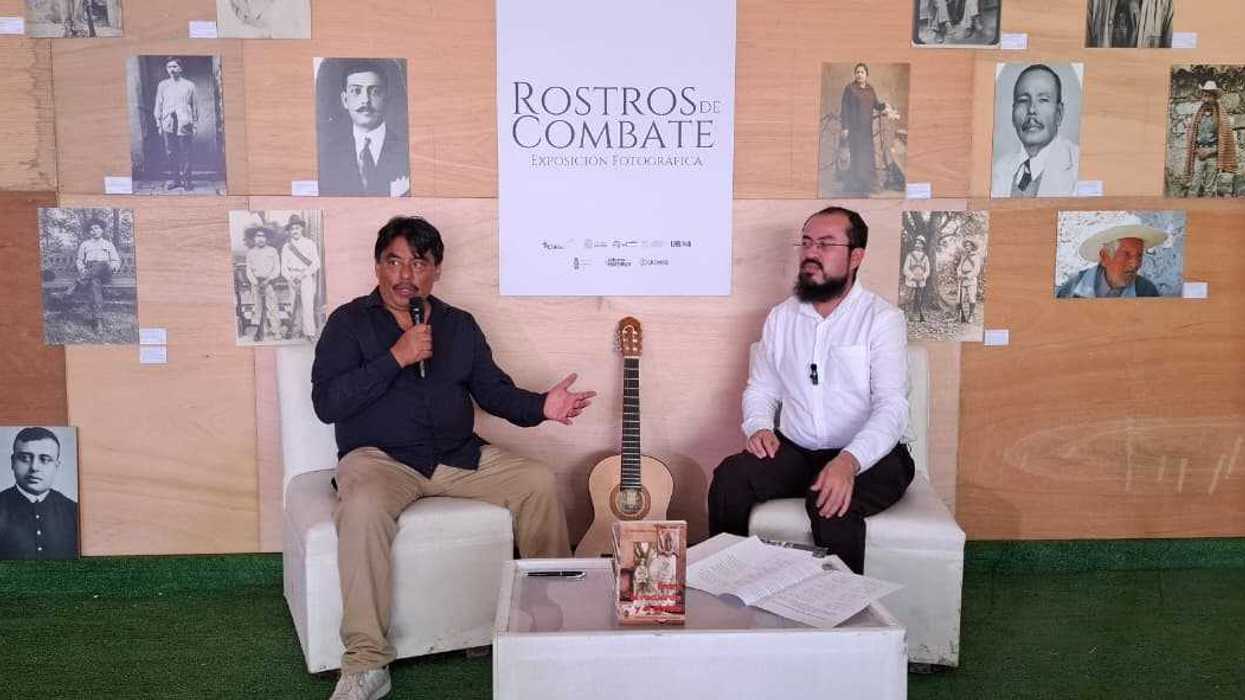 Dos hombres conversan en sillones blancos durante la presentación de un libro, con una guitarra al lado