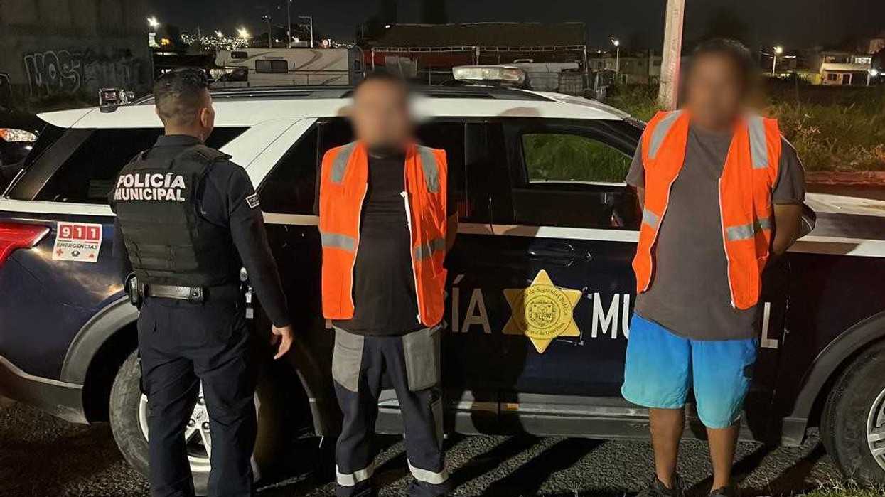 Dos hombres detenidos por daños a la carpeta asfáltica en la colonia Satélite, con chalecos naranjas, junto a una patrulla y un policía municipal