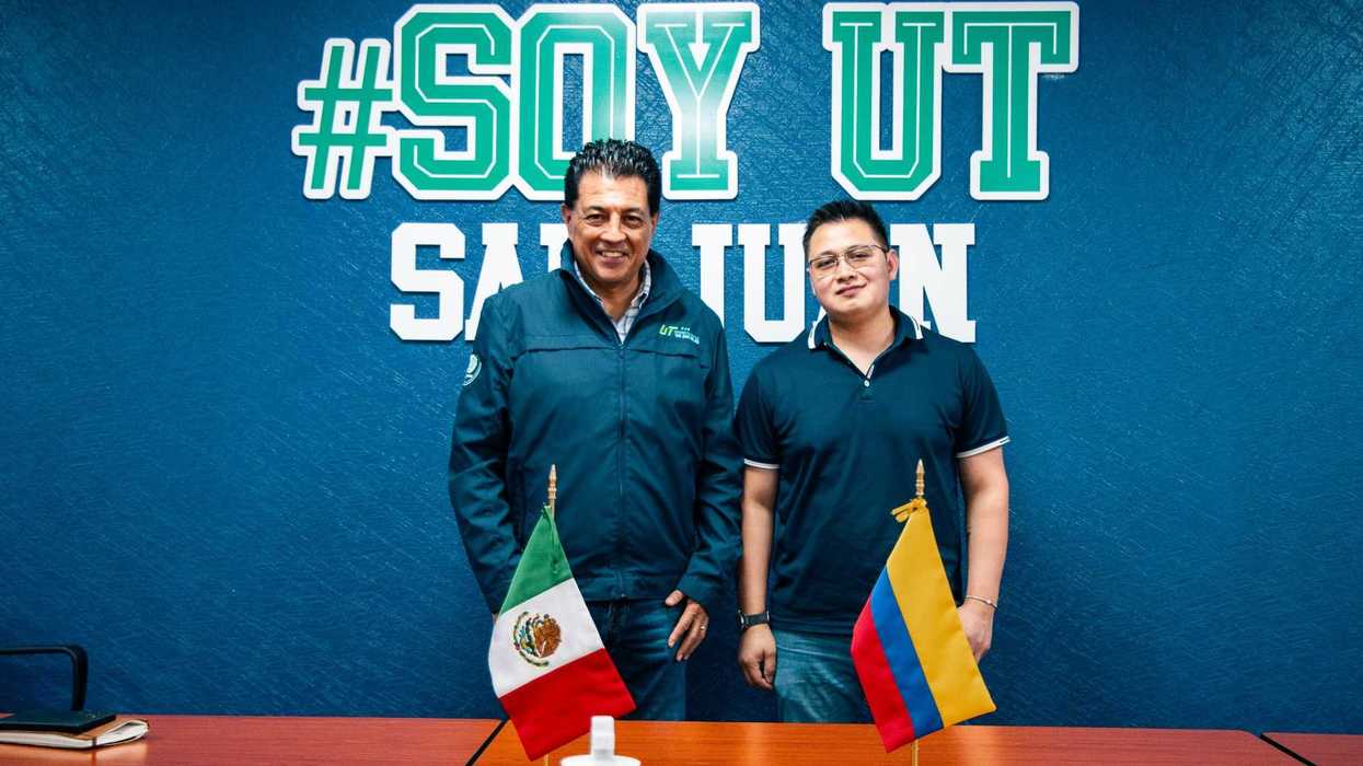 Dos hombres posan frente al muro con el logotipo “#Soy UT San Juan”, con banderas de México y Colombia sobre la mesa