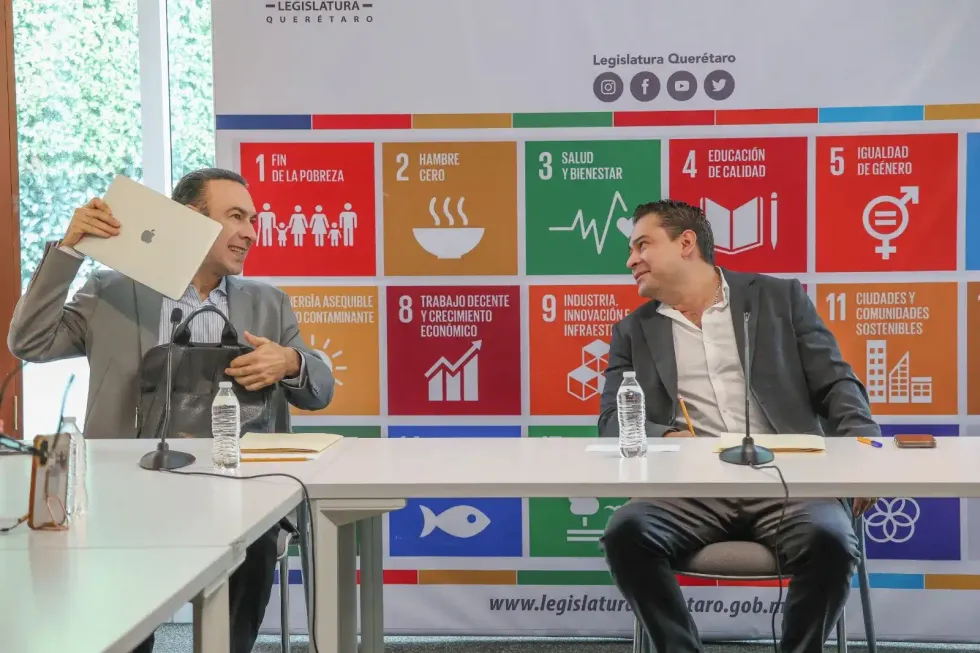 Dos integrantes de la comisi\u00f3n conversan en la mesa de sesi\u00f3n frente al banner de la Agenda 2030.
