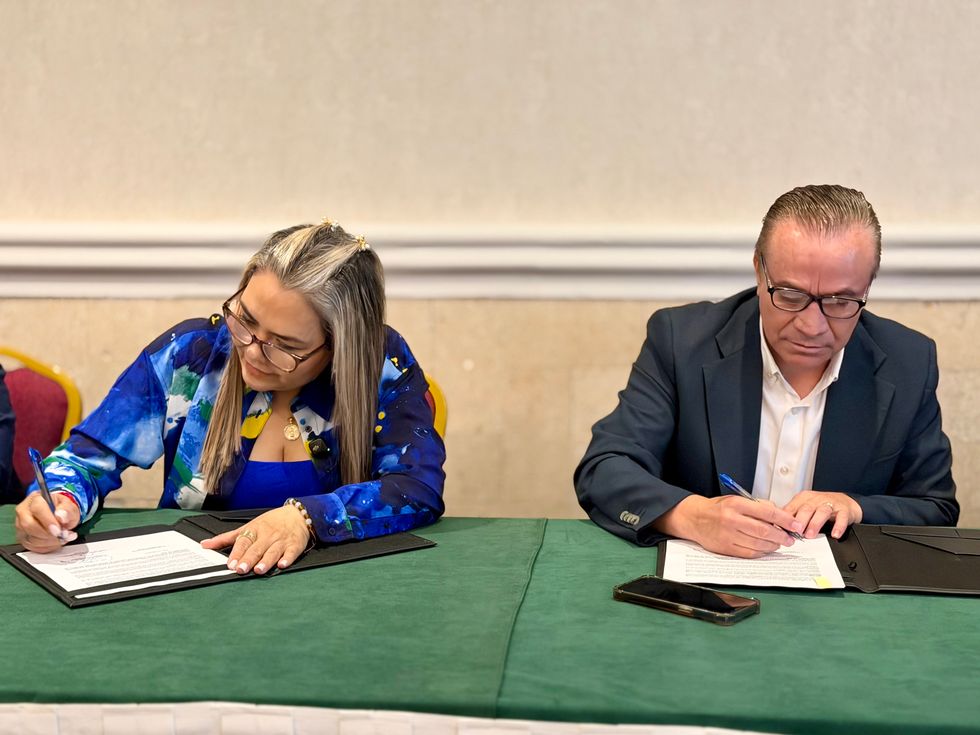 Dos personas firman documentos sobre una mesa durante la firma de un convenio.