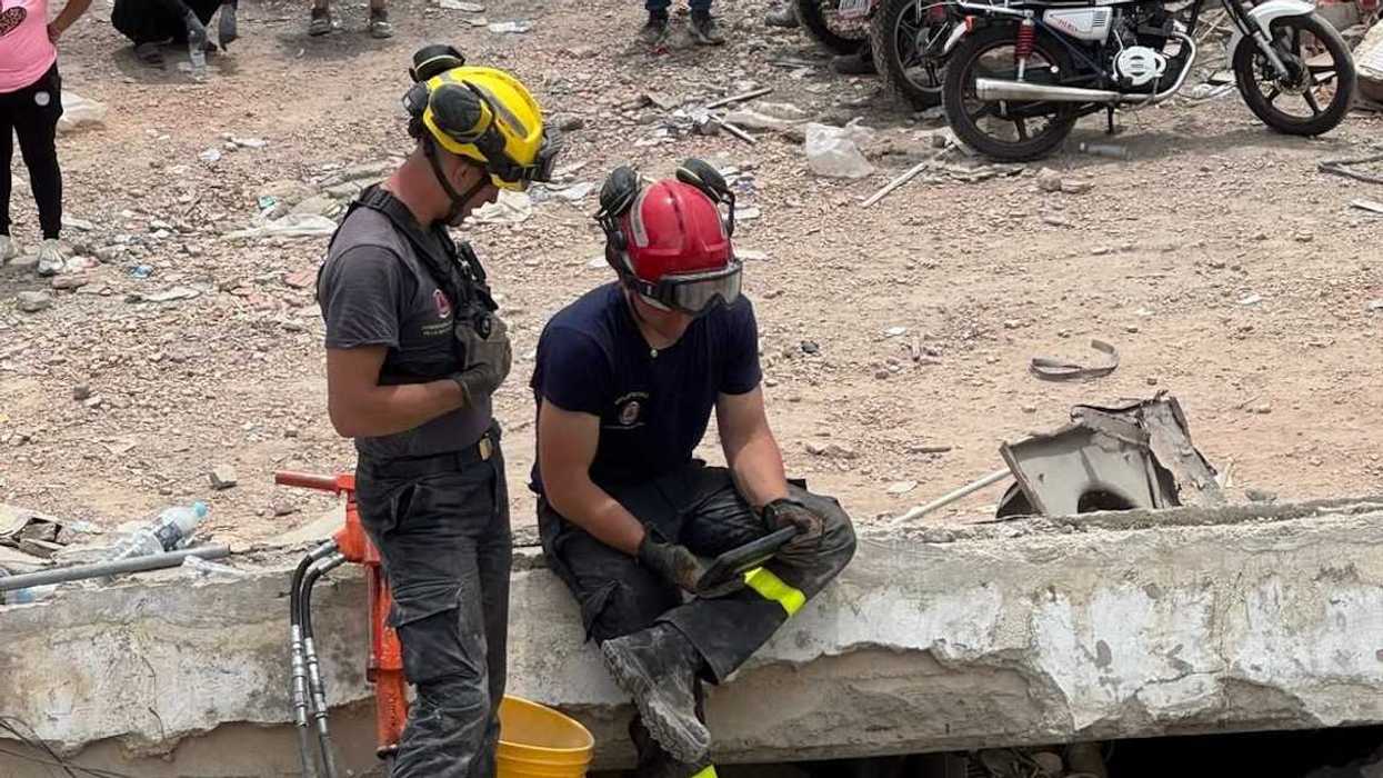 Dos rescatistas con casco descansan sobre los escombros de un edificio colapsado mientras vecinos observan.