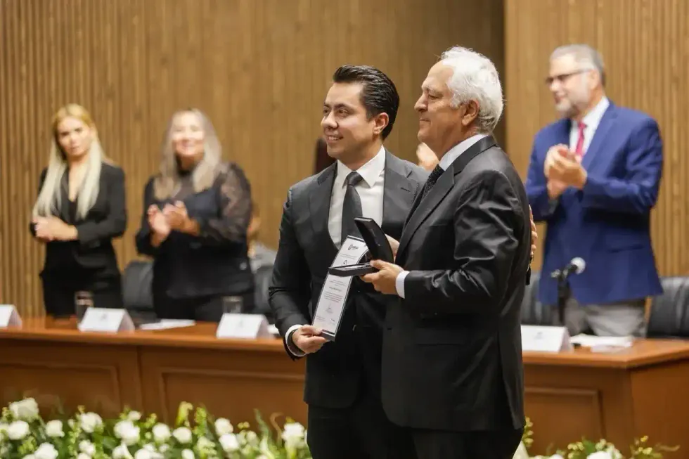 El presidente municipal entrega un reconocimiento enmarcado y una medalla a un hombre de cabello blanco, mientras integrantes del pres\u00eddium aplauden.