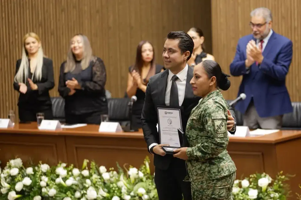 El presidente municipal entrega un reconocimiento y una medalla a una integrante del Ej\u00e9rcito Mexicano, de uniforme, ante el pres\u00eddium.