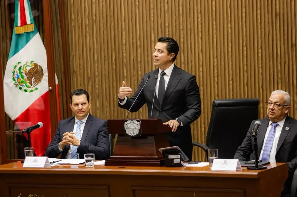 El presidente municipal habla de pie en el pres\u00eddium del sal\u00f3n de Cabildo, flanqueado por dos funcionarios sentados.