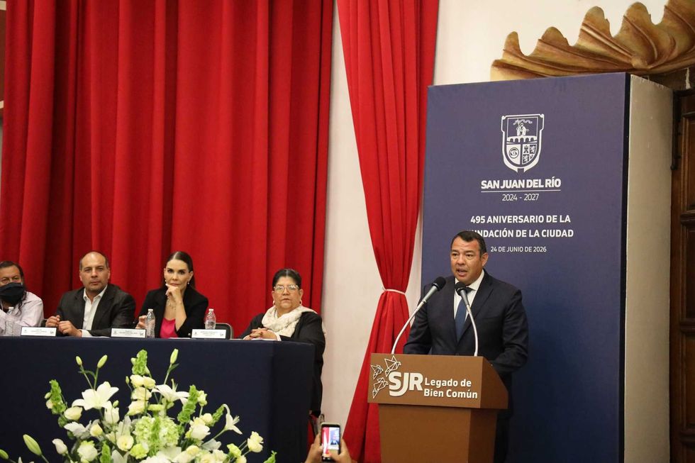 El presidente municipal Roberto Carlos Cabrera Valencia habla en el podio con el panel institucional del 495 aniversario visible y cuatro integrantes del cabildo al fondo en el Foro San Juan
