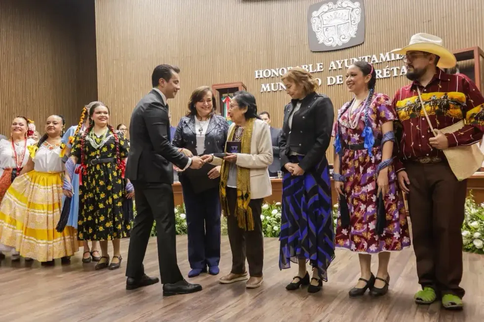 El presidente municipal saluda a una mujer que sostiene un reconocimiento, acompa\u00f1ada por bailarines con trajes tradicionales de distintas regiones.