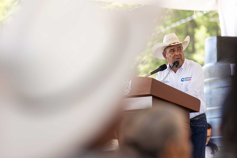 El secretario de Desarrollo Agropecuario, Rosendo Anaya Aguilar, habla en el podio durante el arranque del programa Apoyo al Campo Queretano; al fondo, tinas de captaci\u00f3n de agua entre los implementos entregados.
