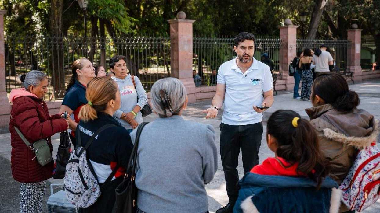 El senador Agustín Dorantes Lámbarri, con playera blanca, muestra un volante azul a una mujer con sombrero de paja frente a una reja verde; imagen ligada a la iniciativa ciudadana Ley Reembolso.
