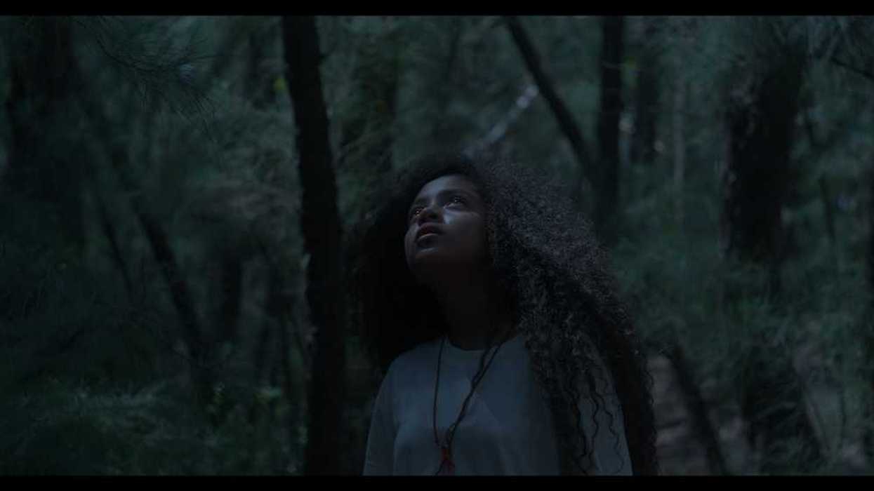 Escena de la película en un bosque en penumbra: una joven de cabello largo y rizado mira hacia arriba