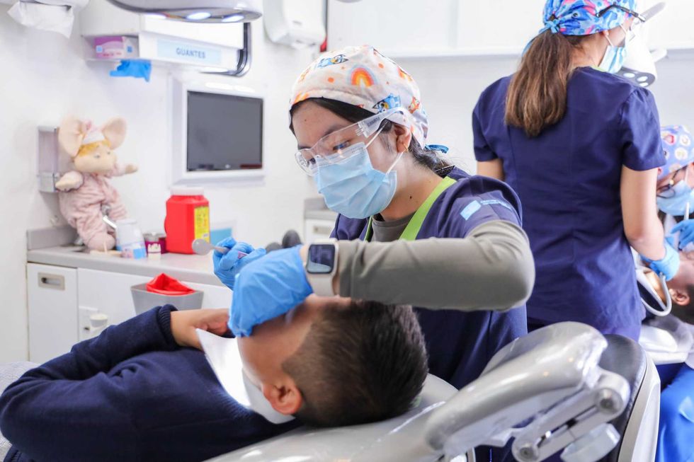 Estudiante de odontolog\u00eda con cubrebocas y lentes de protecci\u00f3n revisa la boca de un menor recostado en el sill\u00f3n de la unidad dental m\u00f3vil.