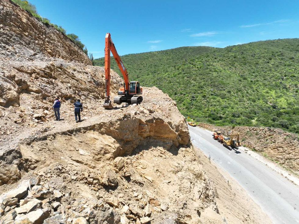 Excavadora naranja trabaja en la parte alta del talud sobre la Carretera Estatal 100, con dos trabajadores de la CEI cerca de la m\u00e1quina