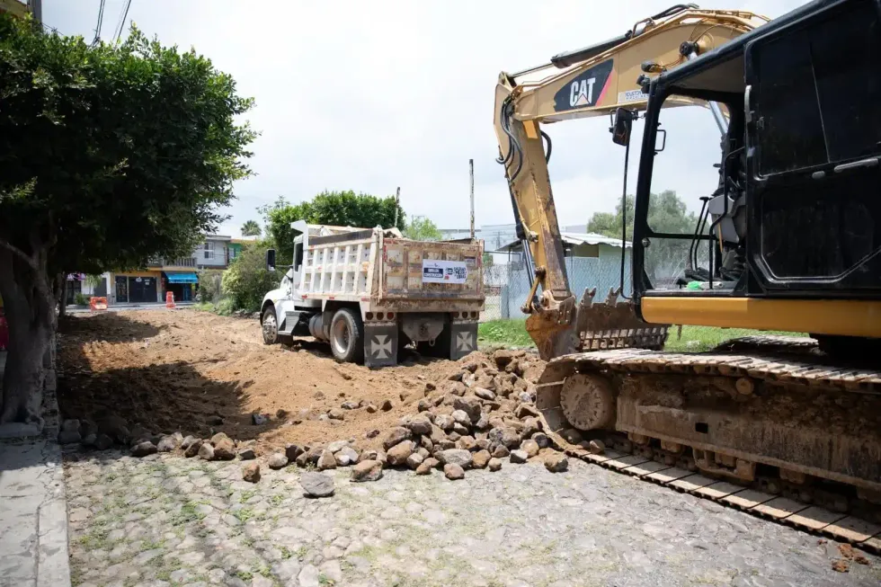 Excavadora y cami\u00f3n de volteo trabajan en el retiro de material sobre una calle empedrada