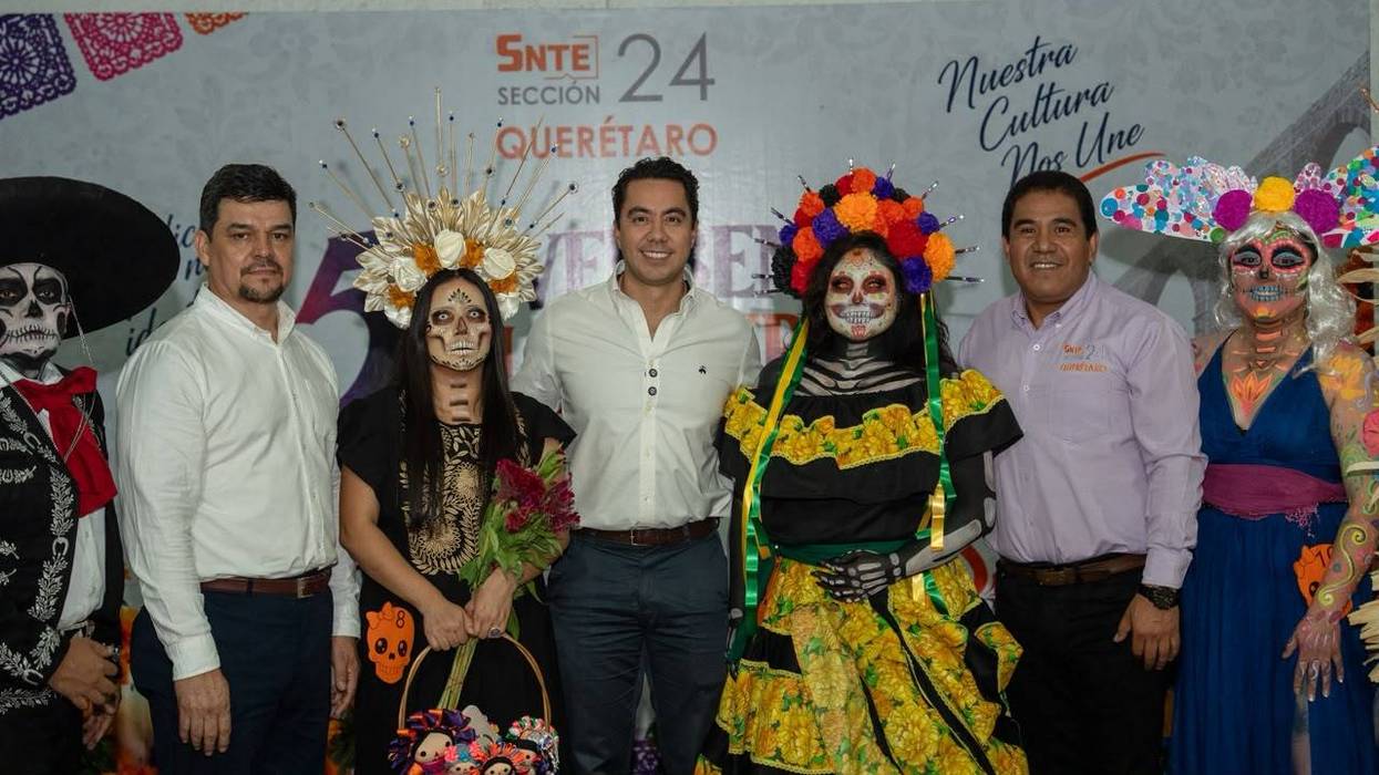 Felifer Macías posa sonriente junto a integrantes del SNTE Sección 24 con catrinas y coronas de cempasúchil, frente a un fondo de la Verbena Magisterial.