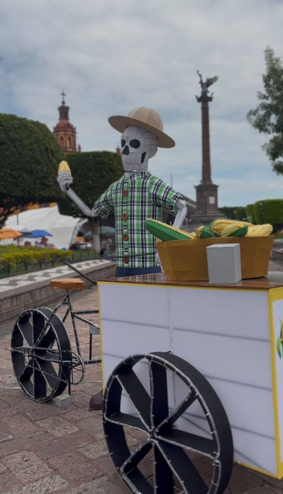 Figura de calavera con sombrero y camisa a cuadros sostiene un elote junto a un carrito de venta, en una plaza del centro de San Juan del R\u00edo
