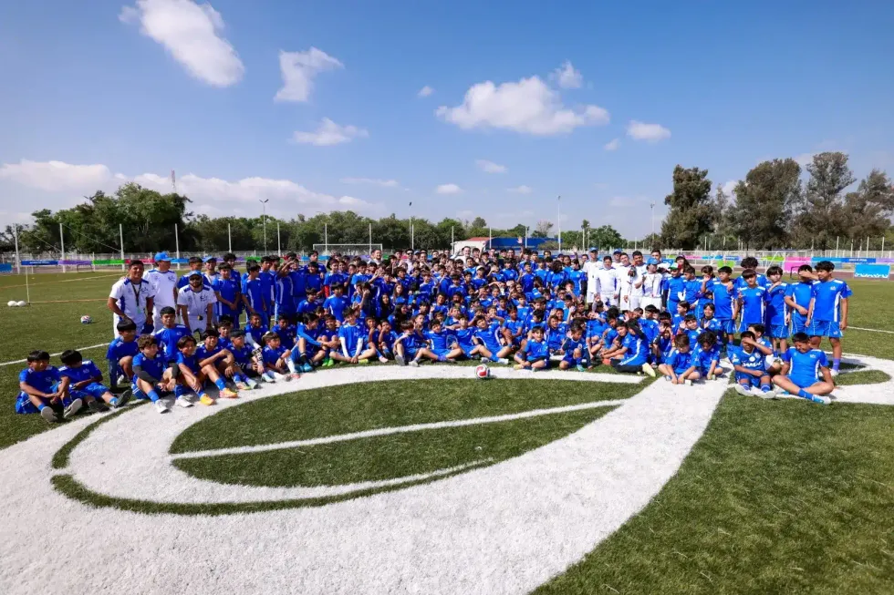Foto grupal de m\u00e1s de ciento cincuenta participantes de la Cl\u00ednica de F\u00fatbol INDEREQ 2026, vestidos con uniformes azules, posando en la cancha de pasto sint\u00e9tico de la unidad deportiva Plutarco El\u00edas Calles de Quer\u00e9taro.