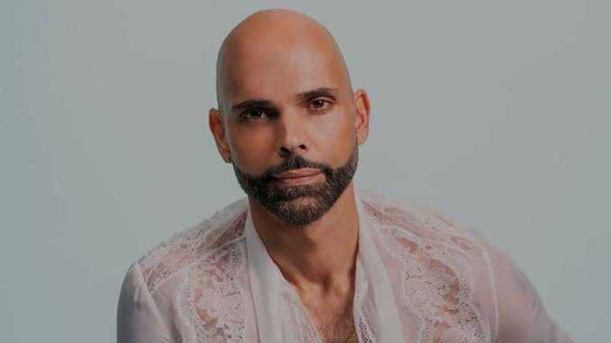 Hombre calvo y con barba, con blusa blanca de encaje translúcido, frente a un fondo gris