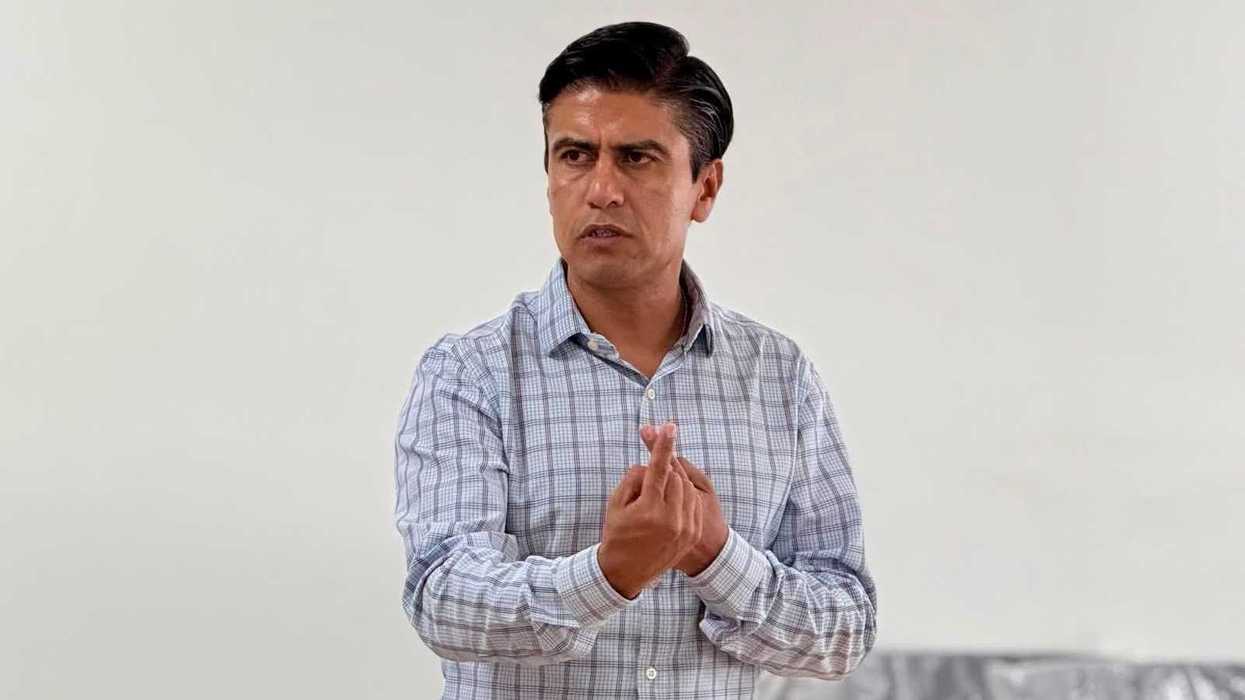 Hombre con camisa a cuadros haciendo un gesto con las manos durante una actividad interna, con sillas cubiertas de plástico al fondo.