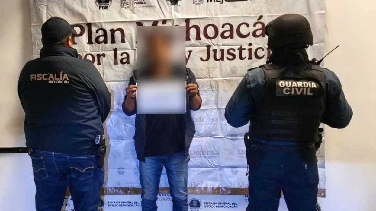 Hombre con el rostro cubierto es custodiado por un agente de la Fiscalía y un elemento de la Guardia Civil frente a una lona de "Plan Michoacán por la Paz y Justicia", en Ciudad Hidalgo.