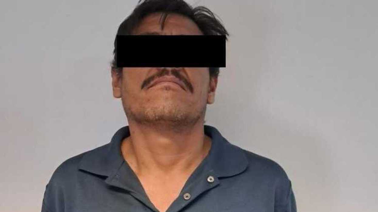 Hombre con playera azul marino, con los ojos cubiertos por una franja negra, de pie frente a un muro claro
