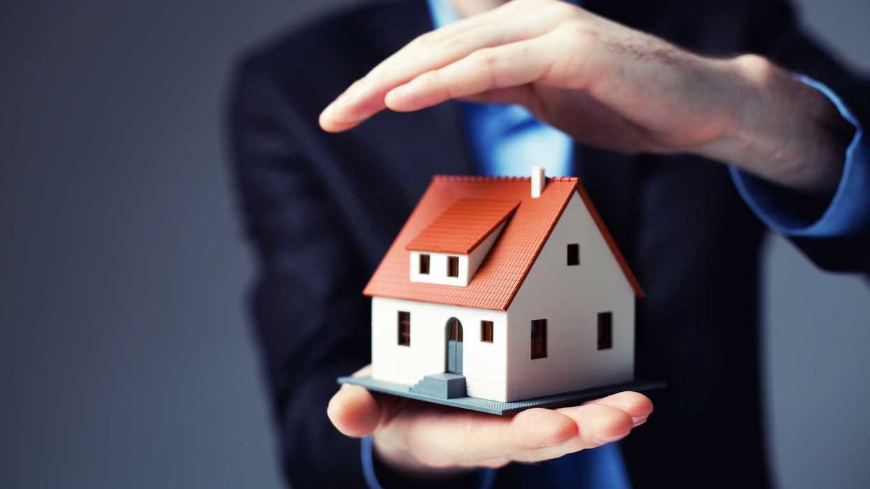 Hombre de traje protege con una mano una casa en miniatura, imagen ilustrativa del seguro de casa habitación.