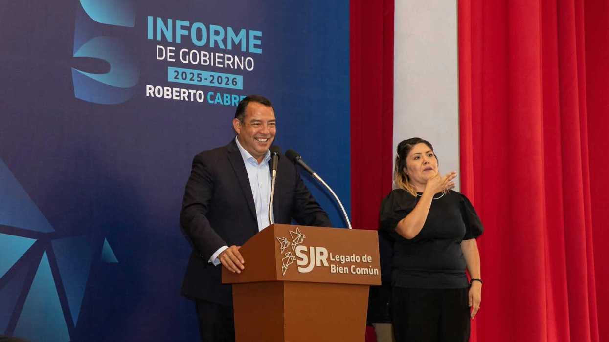 Hombre de traje sonríe frente a un podio junto a una intérprete de lengua de señas, con la lona del Quinto Informe de Gobierno al fondo, en la entrega de escrituras en San Juan del Río.