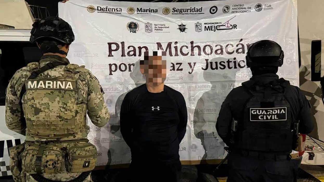 Hombre detenido, con el rostro cubierto por las autoridades, custodiado por elementos de Marina y Guardia Civil frente a lona del Plan Michoacán por la Paz y Justicia