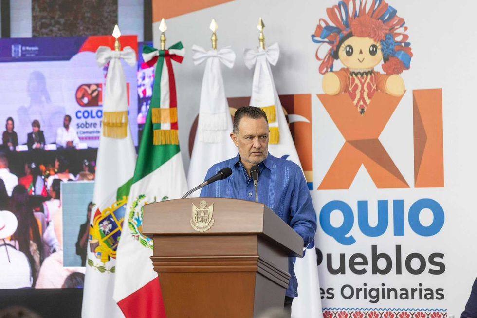 Hombre en guayabera azul estrecha las manos de una mujer ind\u00edgena sentada entre los asistentes