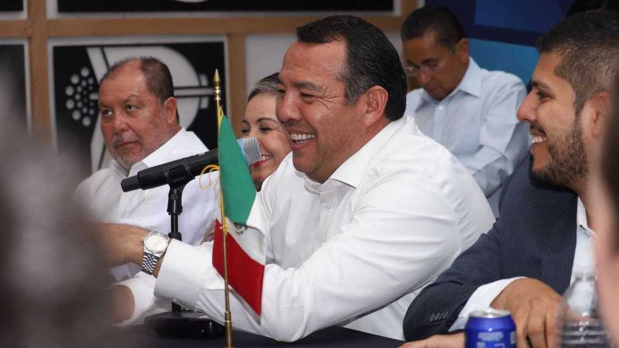 Hombre sonríe al micrófono durante la reunión con agremiados de la CANACO SERVyTUR en San Juan del Río, con una bandera de México sobre la mesa.
