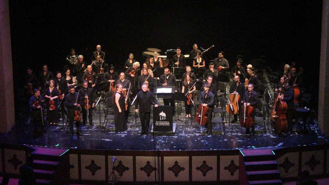 Integrantes de la Orquesta Filarmónica del Estado de Querétaro sobre el escenario durante un concierto, con atril de la agrupación al frente.
