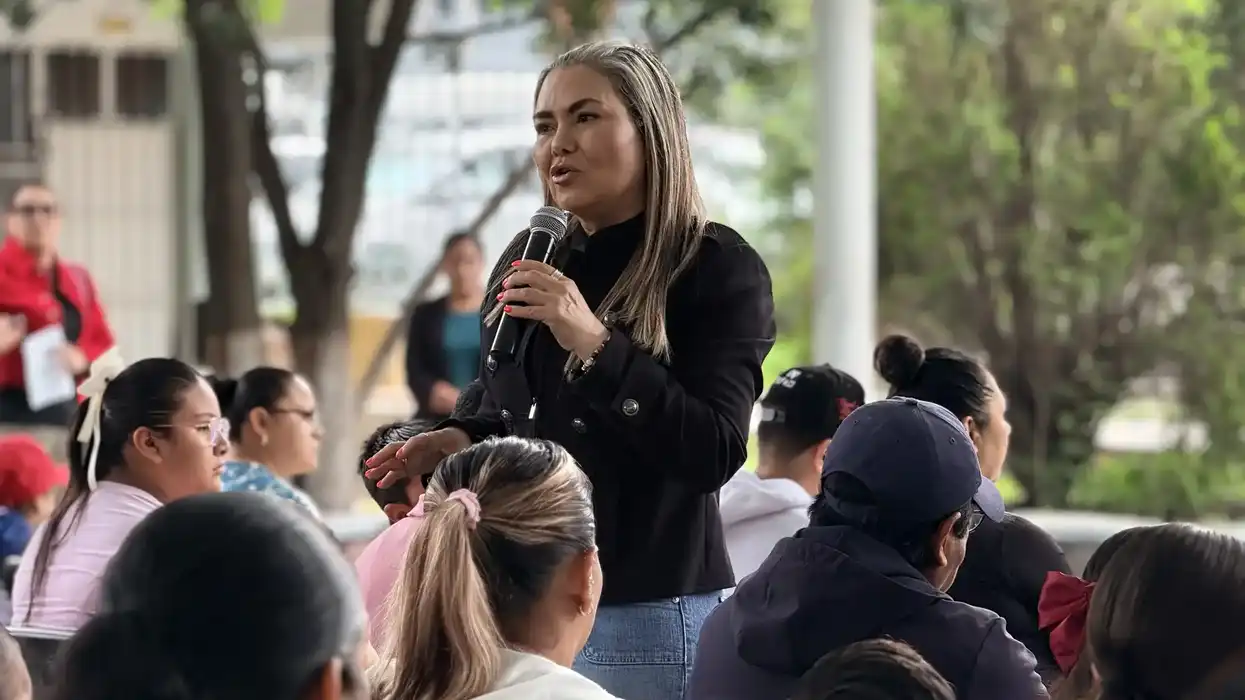 Irene Quintanar habla con micrófono ante padres de familia y estudiantes reunidos al aire libre