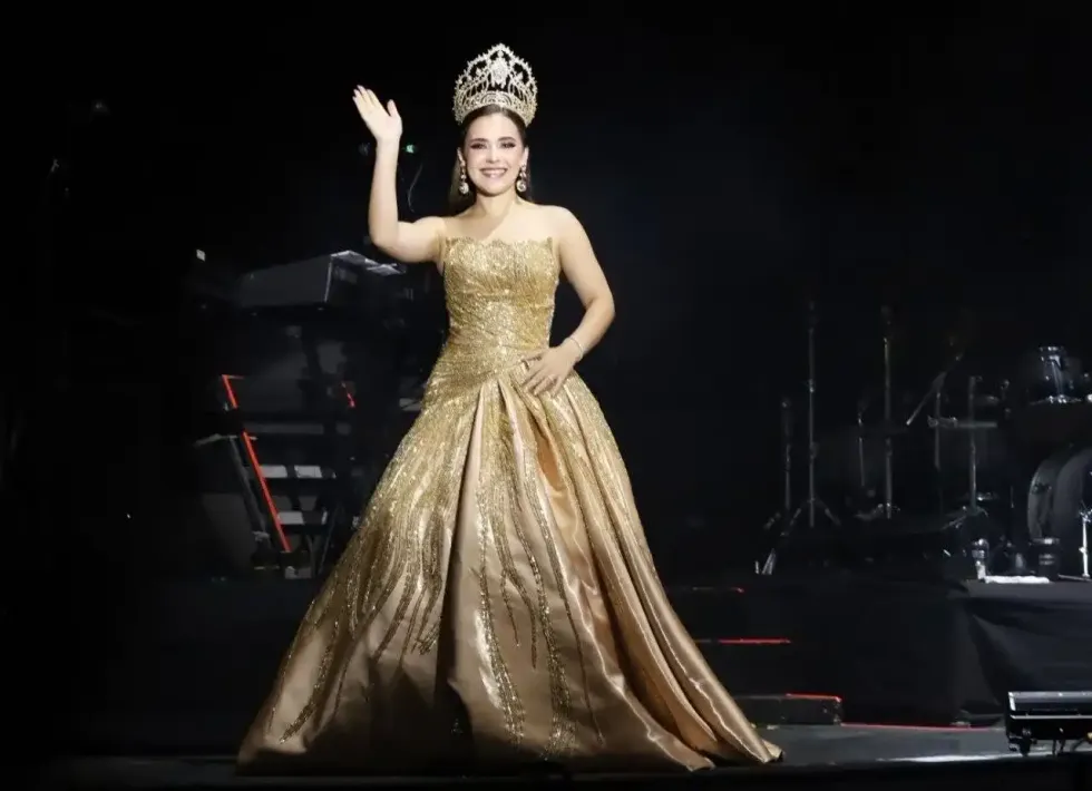 Joven con corona y vestido dorado saluda al p\u00fablico desde el escenario