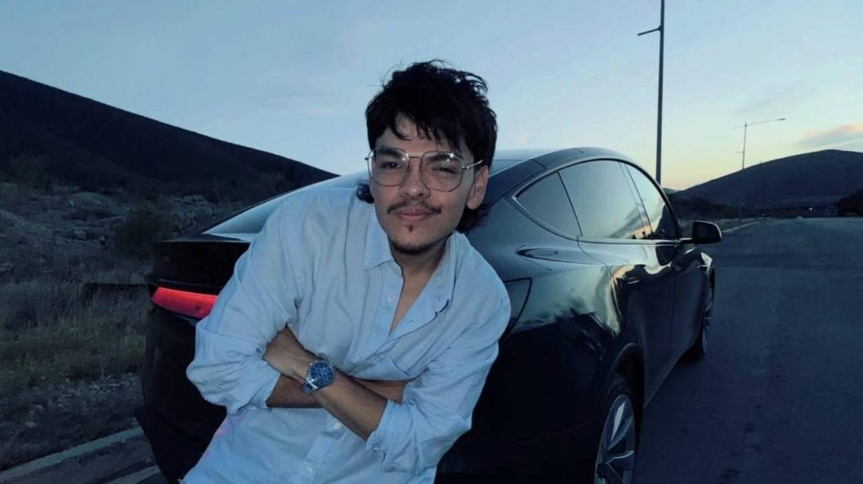 Joven con lentes y camisa clara recargado en un auto oscuro, al anochecer, en una carretera