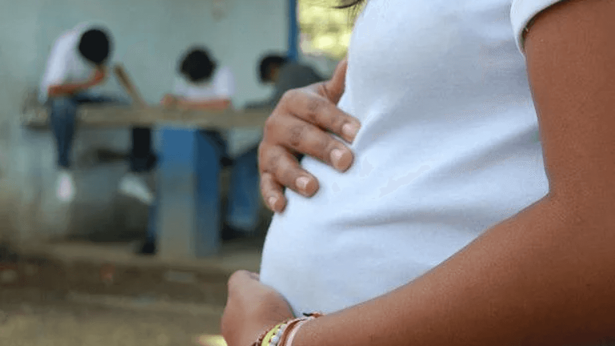Joven embarazada con playera blanca y pulseras de colores sostiene su vientre, con estudiantes al fondo, en referencia al embarazo infantil en México.