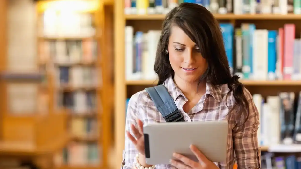 Joven mujer consulta una tablet de pie en una biblioteca, imagen ilustrativa