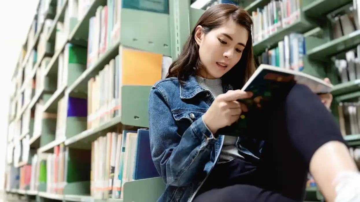 Joven mujer lee un libro sentada en el piso de una biblioteca, entre estantes con libros de colores, imagen ilustrativa